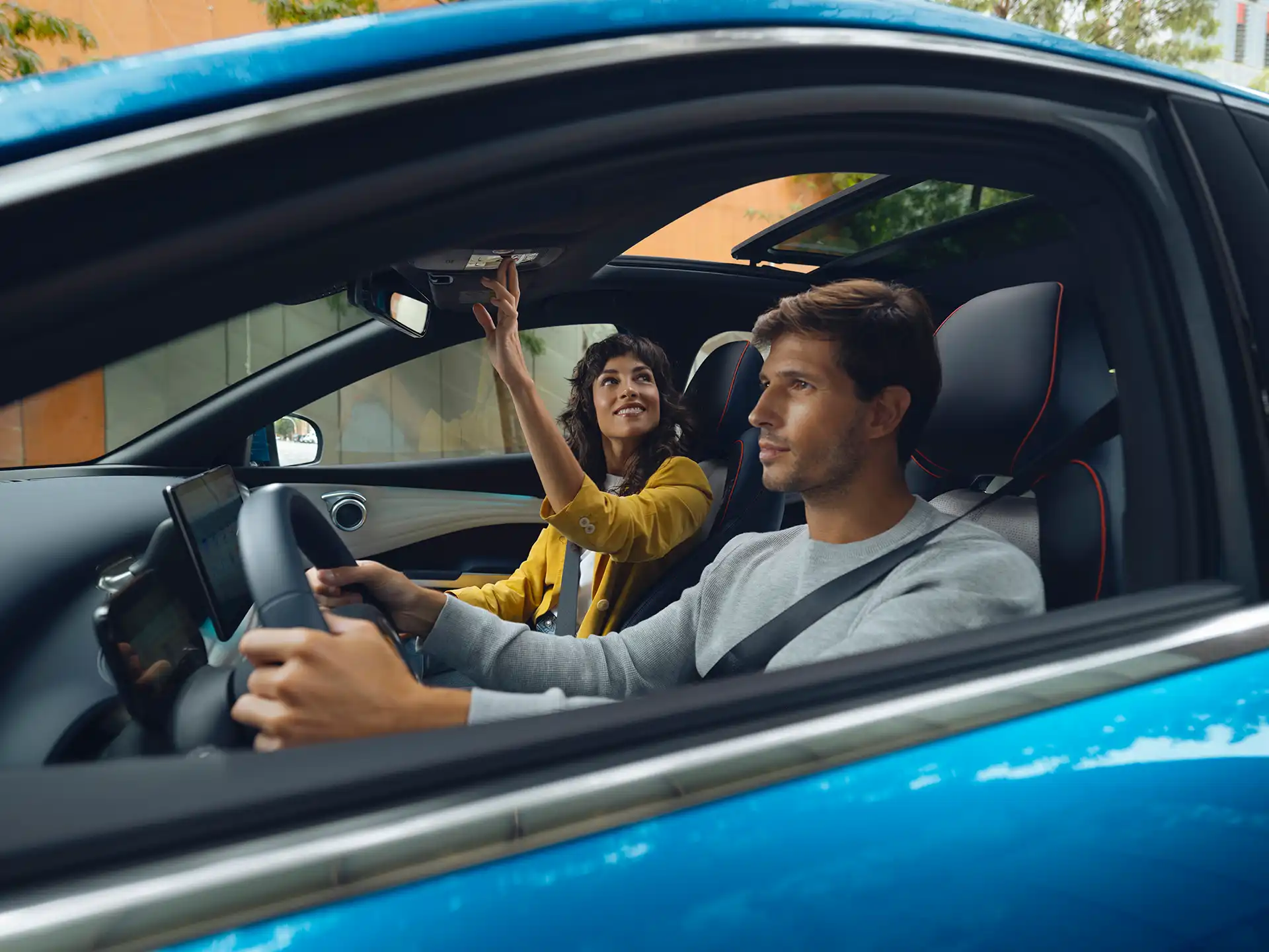 en man en een vrouw zitten in een blauwe elektrische auto; de man rijdt terwijl de vrouw enthousiast naar boven wijst.