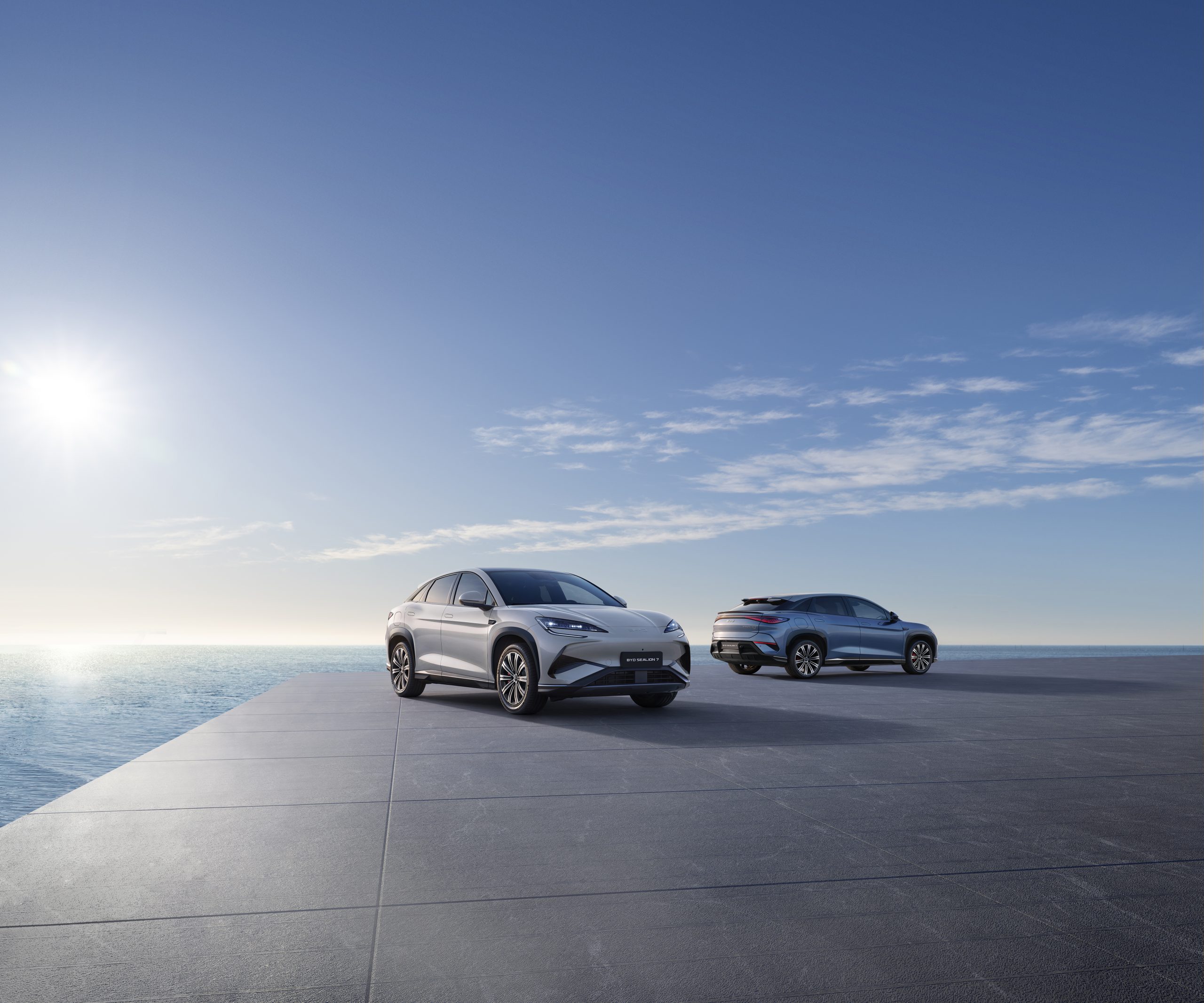 Twee moderne elektrische BYD SUV's geparkeerd op een strak vormgegeven platform met uitzicht op de oceaan, onder een heldere blauwe lucht. De SUV's zijn allebei de BYD SEALION 7.